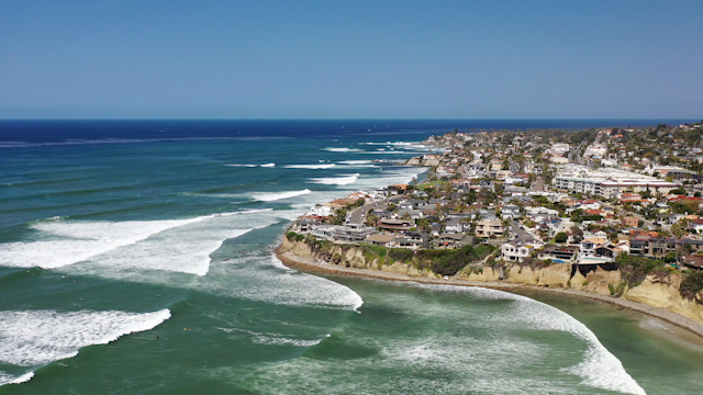 Aerial view of the cliffs, the surf and the ocean in Pacific Beach, with surfers out.