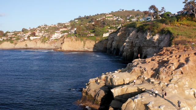 Low aerial view of the cliffs, the ocean and homes at La Jolla Cove, with people walking.