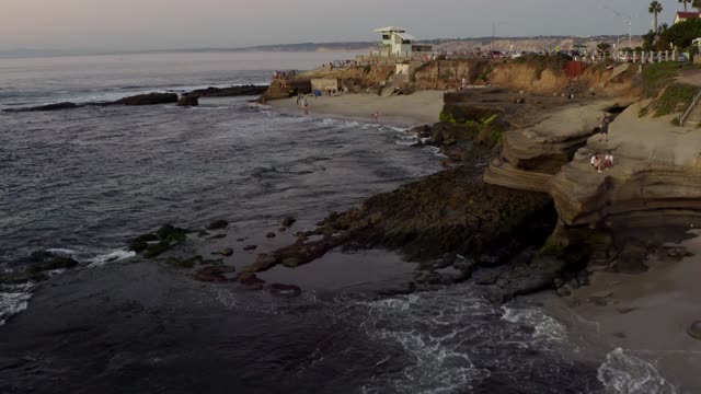 Low aerial view of the cliffs, the ocean and the beach at La Jolla Cove, with people walking.