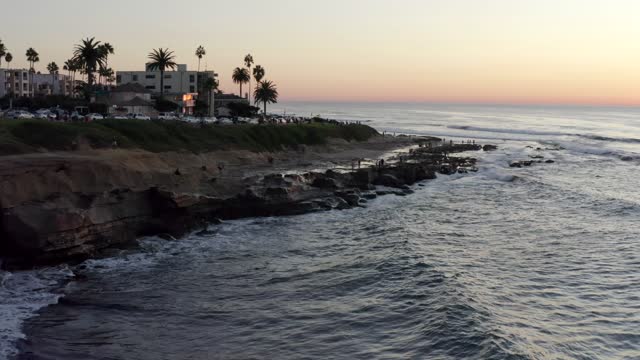 Low aerial view of the cliffs, the surf and palm trees at La Jolla Cove, with people walking.