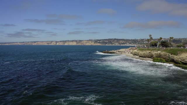 Low aerial view of the cliffs, the surf and palm trees at La Jolla Cove, with people walking.