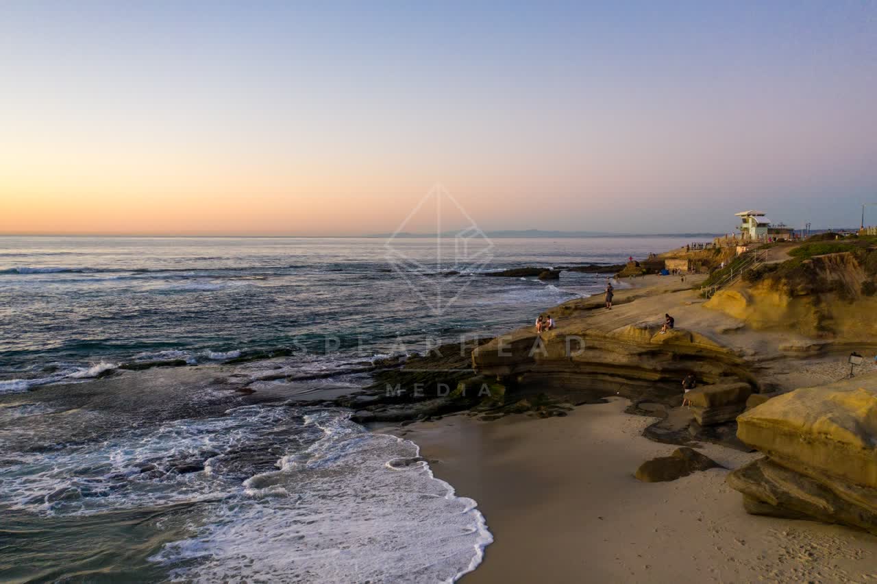 Low aerial view of the cliffs, the surf and the ocean at La Jolla Cove, with people walking.