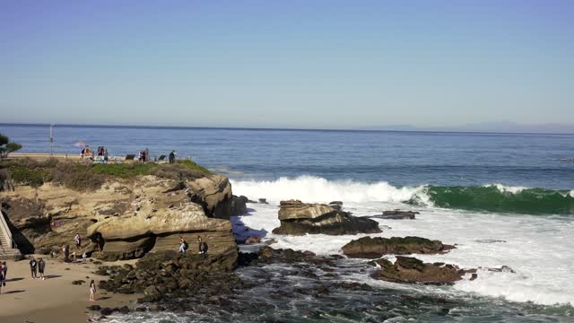 Elevated view of the cliffs, the surf and the ocean at La Jolla Cove, with people walking.