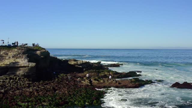 Elevated view of the cliffs, the surf and the ocean at La Jolla Cove, with people walking.