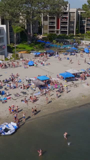 Low aerial view of the beach, palm trees and the bay in Mission Bay, with a crowd gathered.