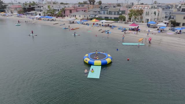 Low aerial view of the beach, boats and the bay in Mission Bay, with a crowd gathered.