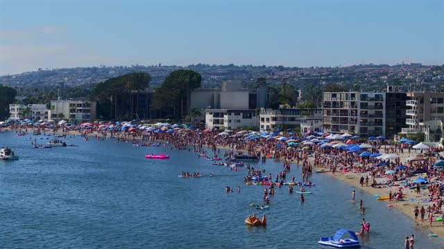 Low aerial view of the beach, boats and the bay in Mission Bay, with a crowd gathered.
