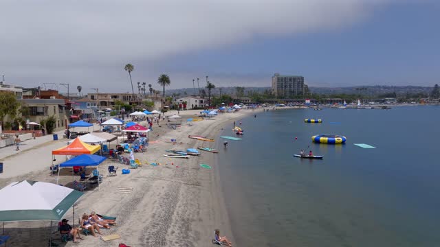 Low aerial view of the beach, boats and palm trees in Mission Bay, with a crowd gathered.