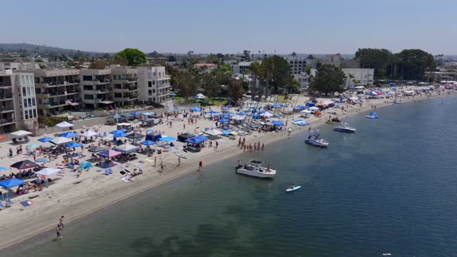 Low aerial view of the beach, boats and the bay in Mission Bay, with a crowd gathered.