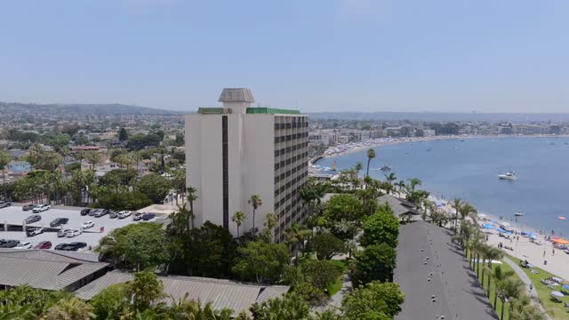 Low aerial view of the beach, boats and palm trees in Mission Bay, with a crowd gathered.