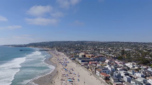Aerial view of the beach, the surf and the pier in Mission Bay, with a crowd gathered.