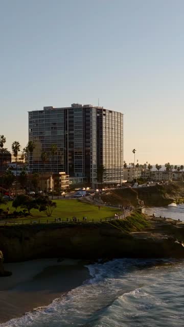 Low aerial view of the cliffs, palm trees and the ocean at La Jolla Cove, with people walking.