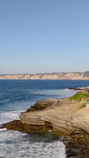 Low aerial view of the cliffs, the surf and the ocean at La Jolla Cove, with people walking.