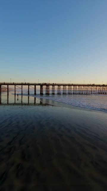 Low aerial view of the beach, the surf and the pier in Oceanside, with people walking.