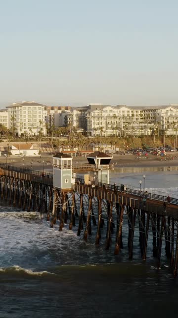 Low aerial view of the beach, the surf and the pier in Oceanside, with people walking.