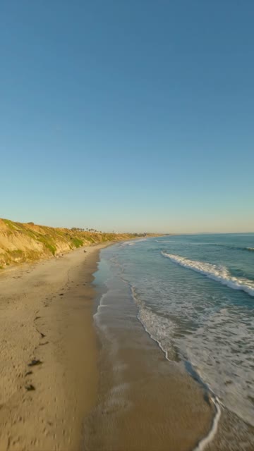 Low aerial view of the beach, the surf and the ocean in Carlsbad, with people walking.
