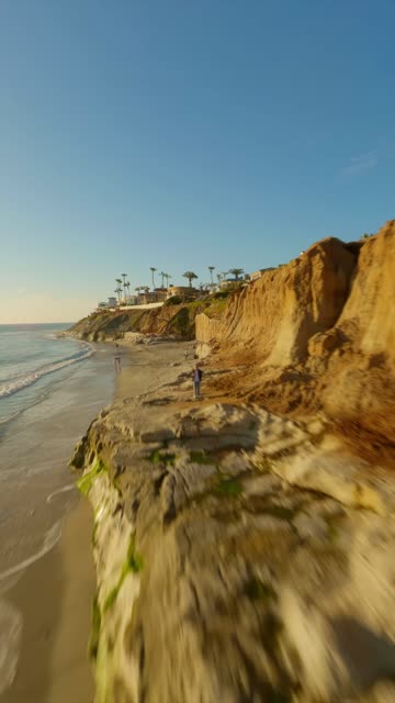 Low aerial view of the cliffs, the surf and palm trees in Carlsbad, with people walking.