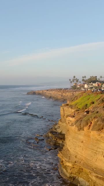 Low aerial view of the cliffs, the surf and palm trees at Sunset Cliffs, with a crowd gathered.