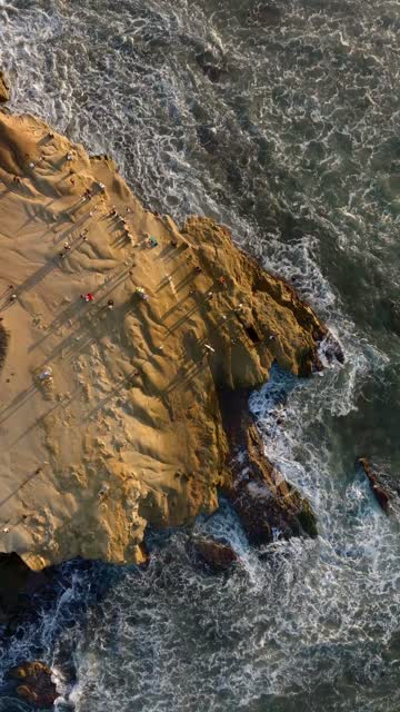 Vertical drone video of King Tides at Sunset Cliffs Natural Park in Point Loma San Diego California during sunset with powerful waves crashing against rugged coastal cliffs and crowds gathering to watch