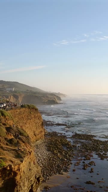 Low aerial view of the cliffs, the surf and the ocean at Sunset Cliffs, with a crowd gathered.