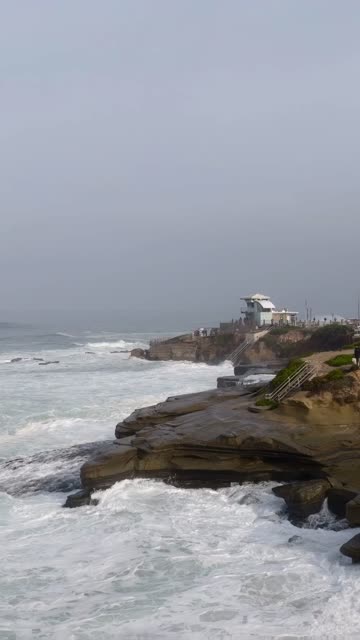 Low aerial view of the cliffs, the surf and the ocean at La Jolla Cove, with people walking.