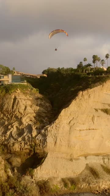 Vertical drone video flying along Black’s Beach in La Jolla California during an extravagant sunset with glowing clouds powerful surf dolphins in motion and paragliders drifting over the bluffs