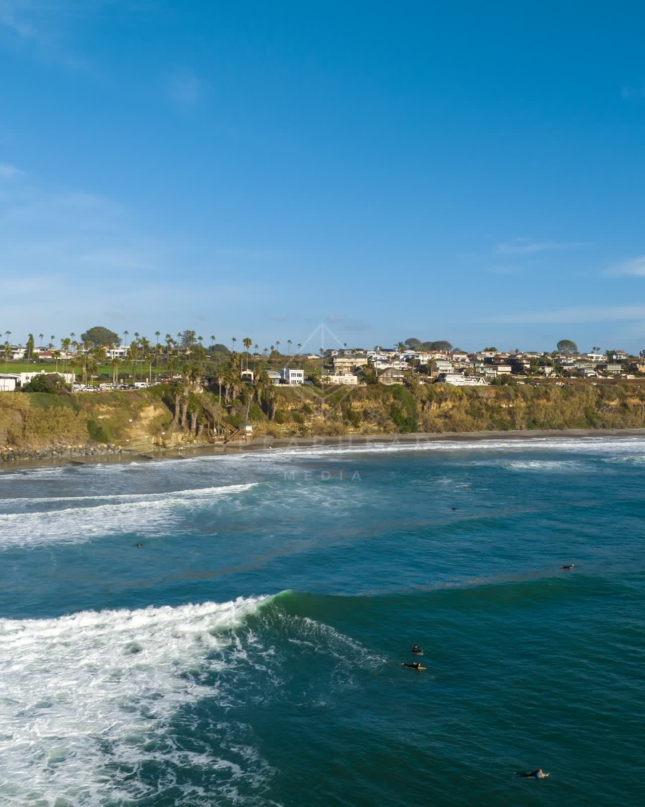 Aerial view of the cliffs, the surf and palm trees at Swamis, with surfers out.
