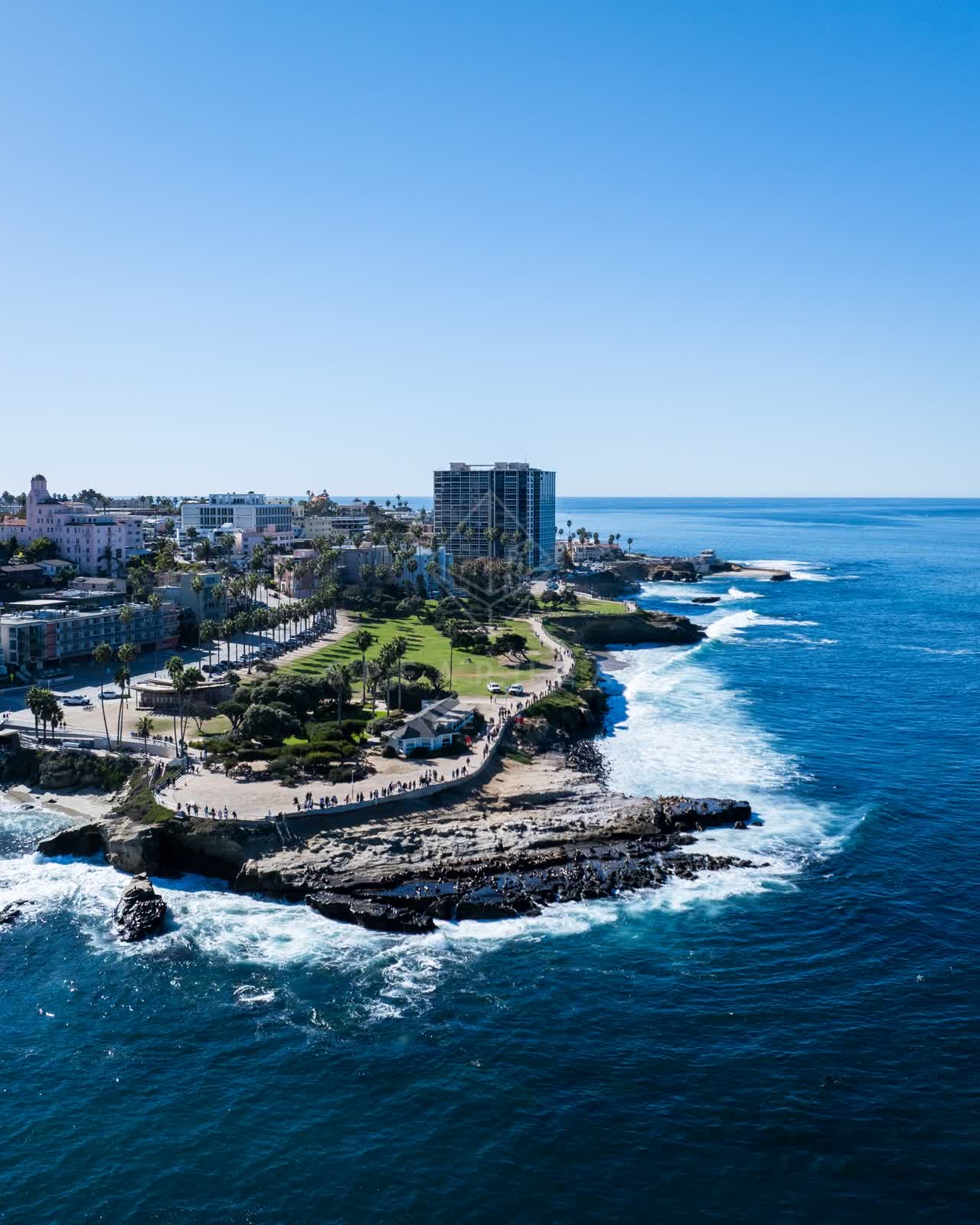 Aerial view of the cliffs, the surf and palm trees at La Jolla Cove, with people walking.
