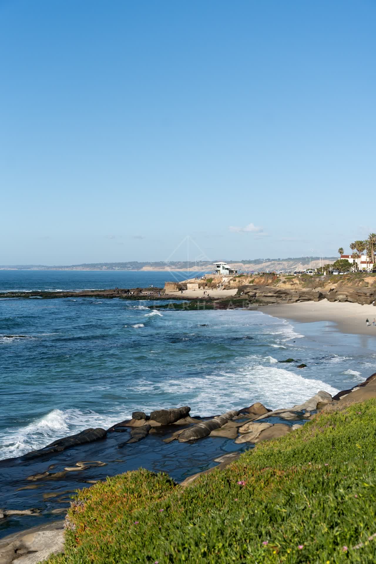 Elevated view of the cliffs, the surf and palm trees at La Jolla Cove, with people walking.