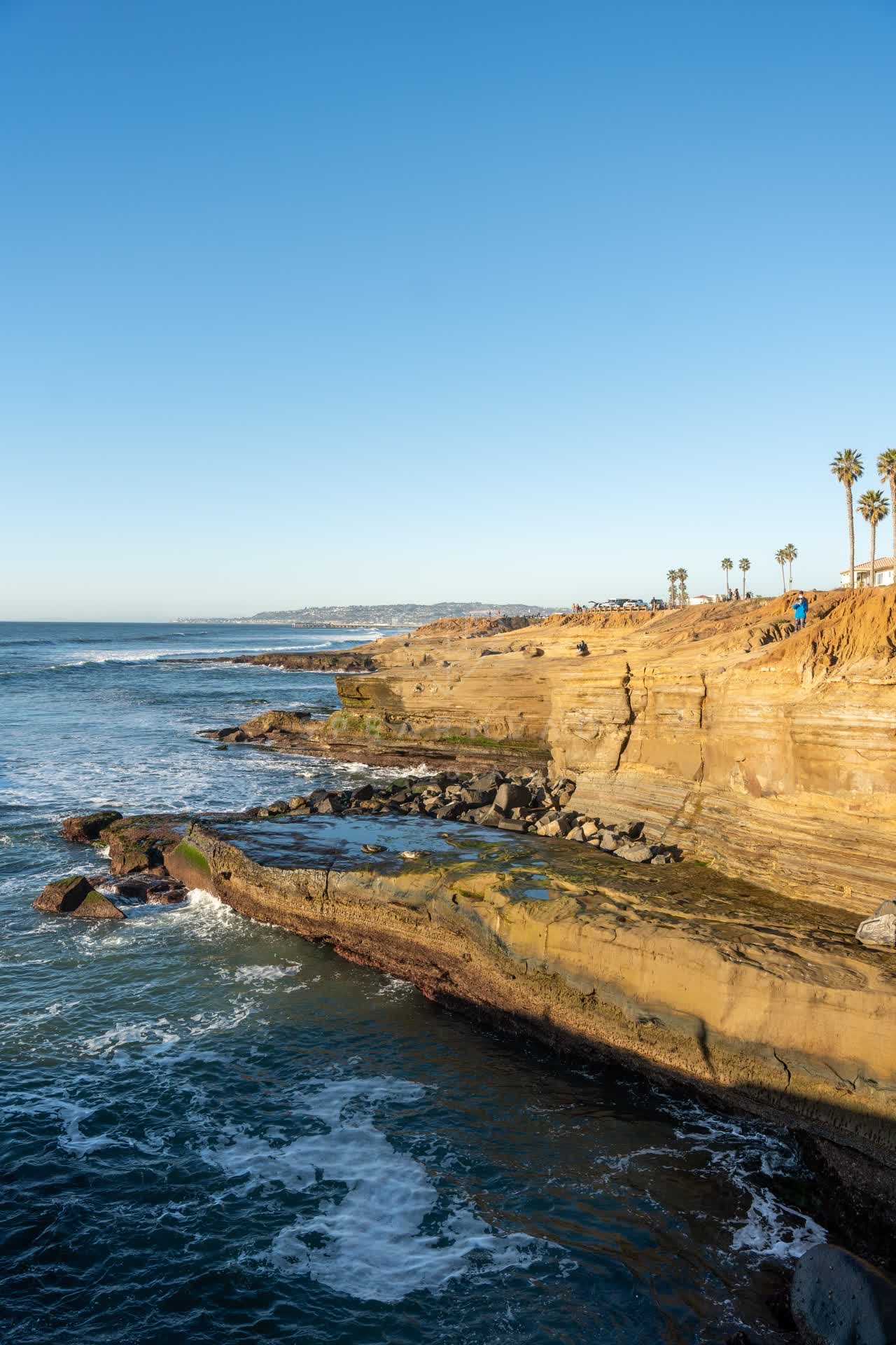Elevated view of the cliffs, the surf and palm trees at Sunset Cliffs, with people walking.