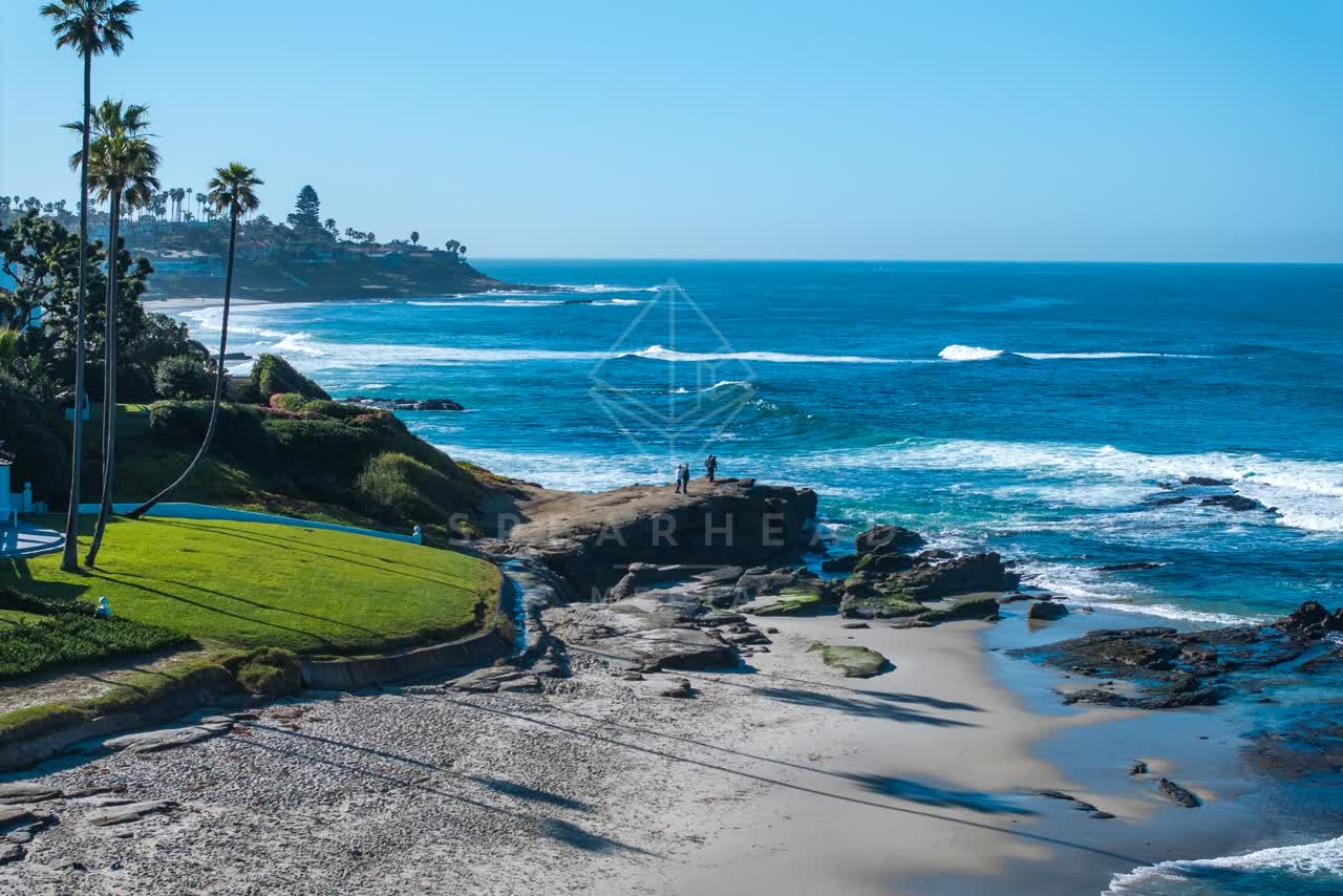 Low aerial view of the cliffs, the surf and palm trees at Windansea, with people walking.