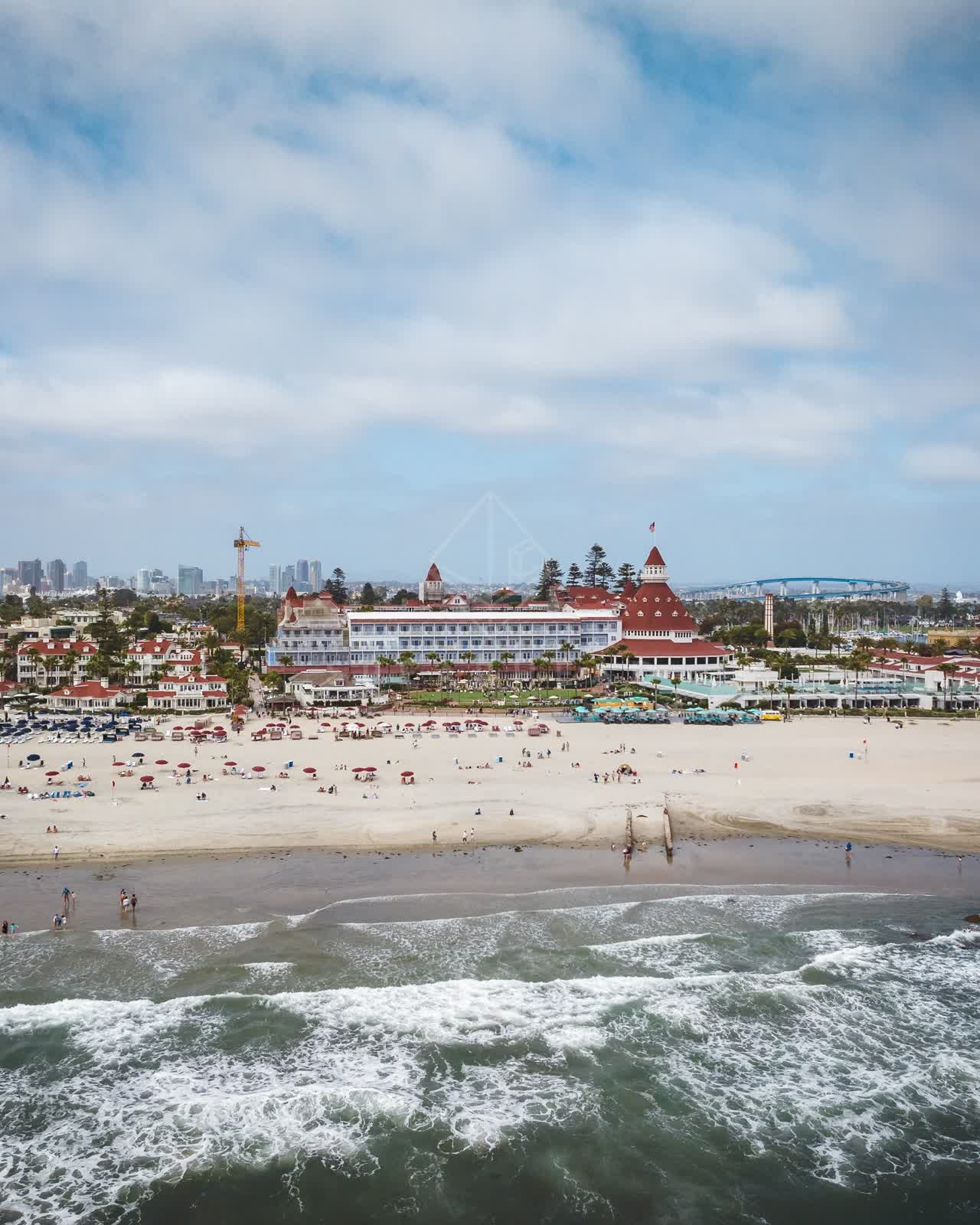 Aerial view of the beach, the surf and the bridge at Coronado Beach, with people walking.