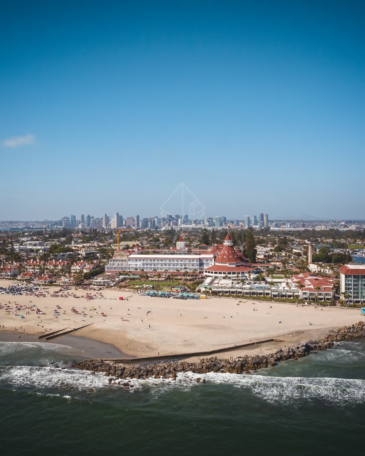Aerial view of the beach, the surf and the skyline at Coronado Beach, with a crowd gathered.