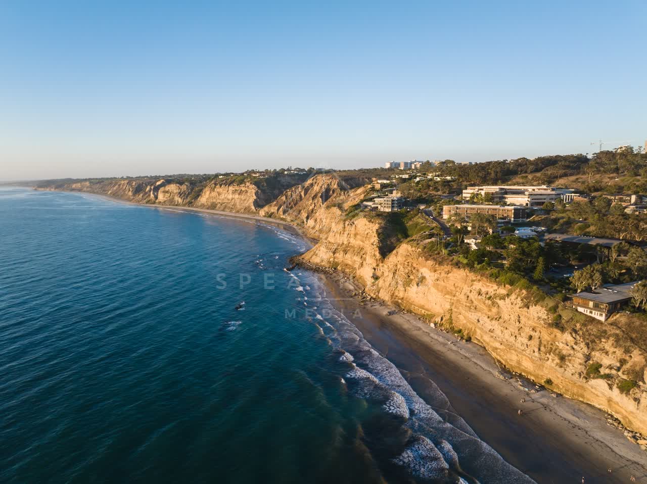 Aerial view of the cliffs, the surf and the ocean at Black’s Beach, with surfers out.