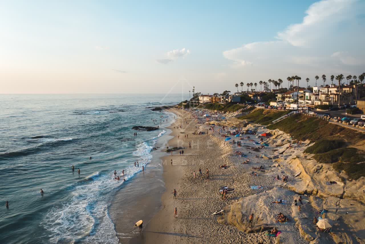Low aerial view of the beach, the surf and palm trees at Windansea, with a crowd gathered.