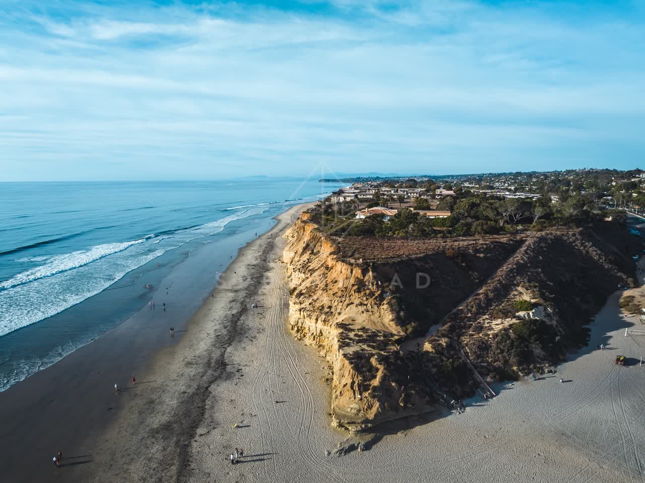Aerial view of the cliffs, the surf and palm trees at Del Mar Dog Beach, with people walking.