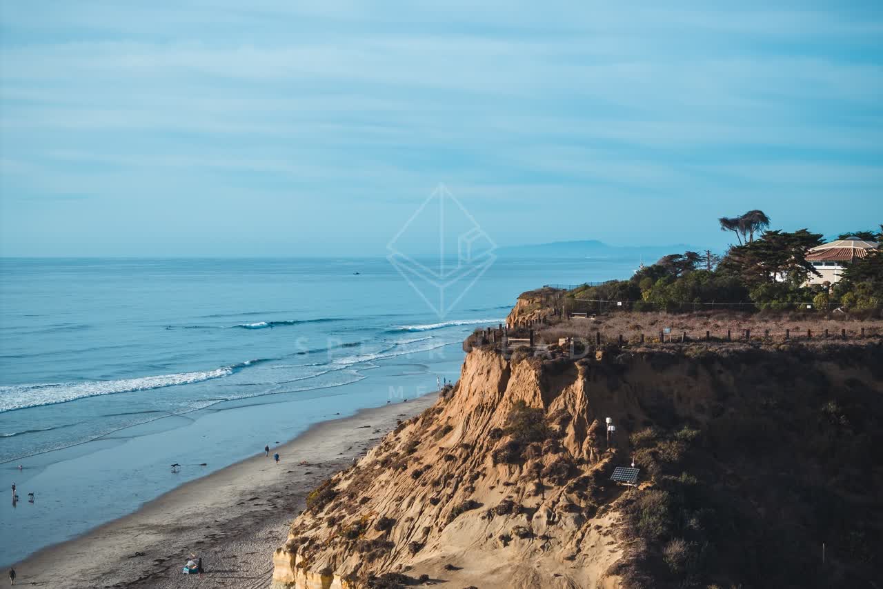 Aerial view of the cliffs, the surf and palm trees at Del Mar Dog Beach, with people walking.