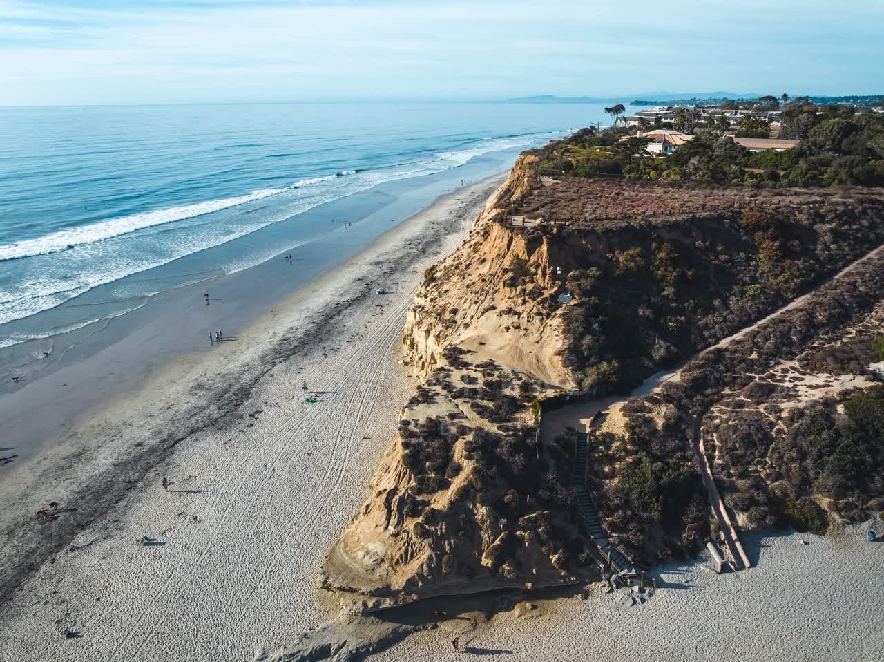 Aerial view of the cliffs, the surf and palm trees at Del Mar Dog Beach, with people walking.