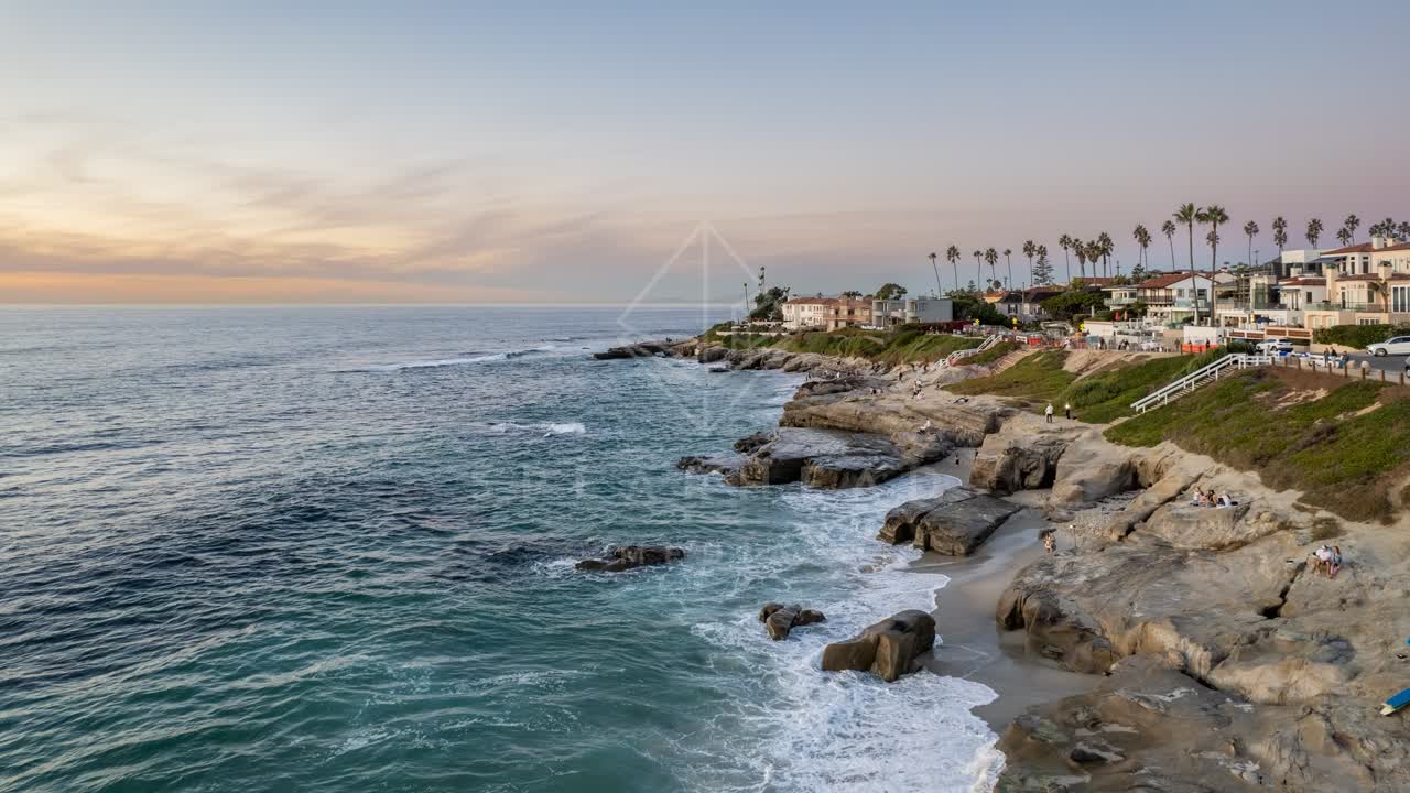 Low aerial view of the cliffs, palm trees and the ocean at Windansea, with people walking.