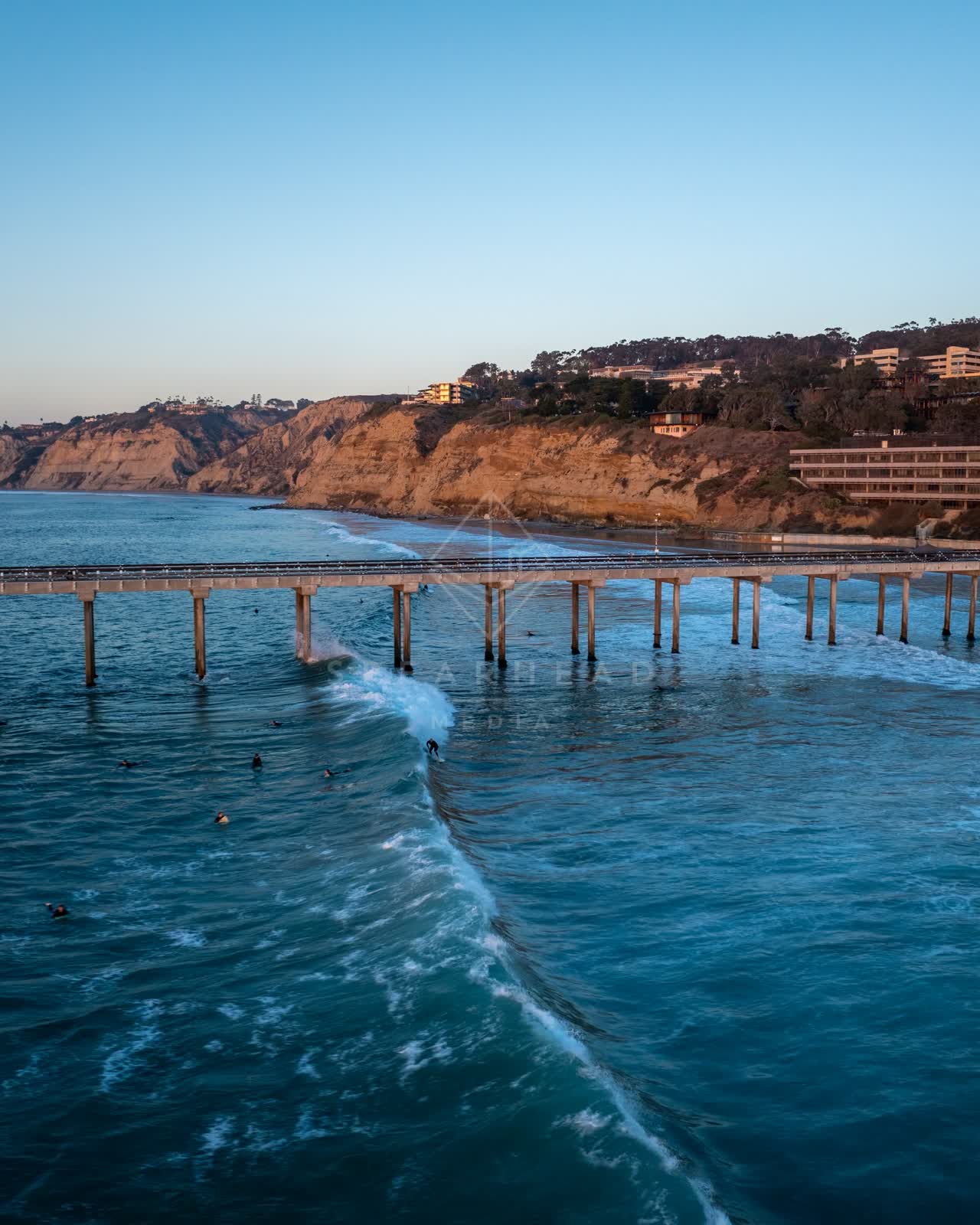 Low aerial view of the beach, the surf and the pier at La Jolla Shores, with surfers out.