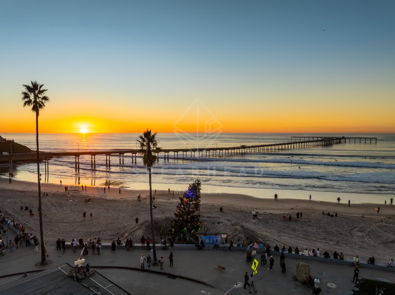 Low aerial view of the beach, the surf and the pier in Ocean Beach, with a crowd gathered.