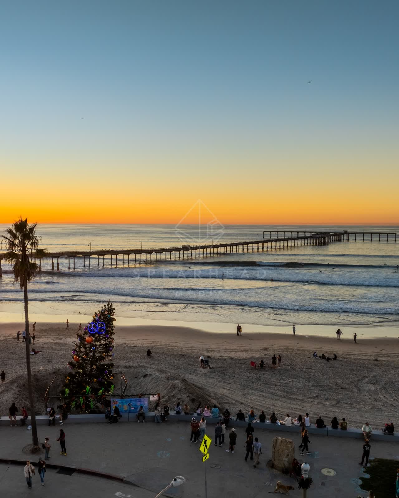 Low aerial view of the beach, the surf and the pier in Ocean Beach, with a crowd gathered.