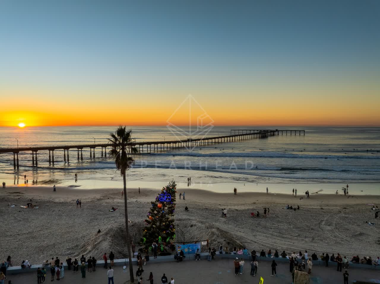 Low aerial view of the beach, the surf and the pier in Ocean Beach, with a crowd gathered.
