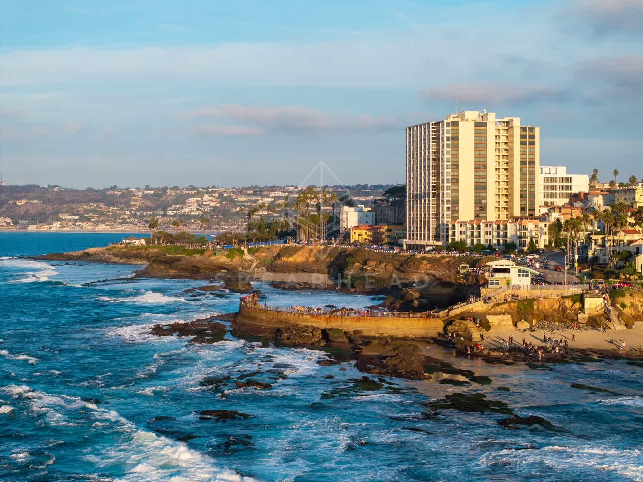 Aerial view of the cliffs, the surf and palm trees at La Jolla Cove, with a crowd gathered.