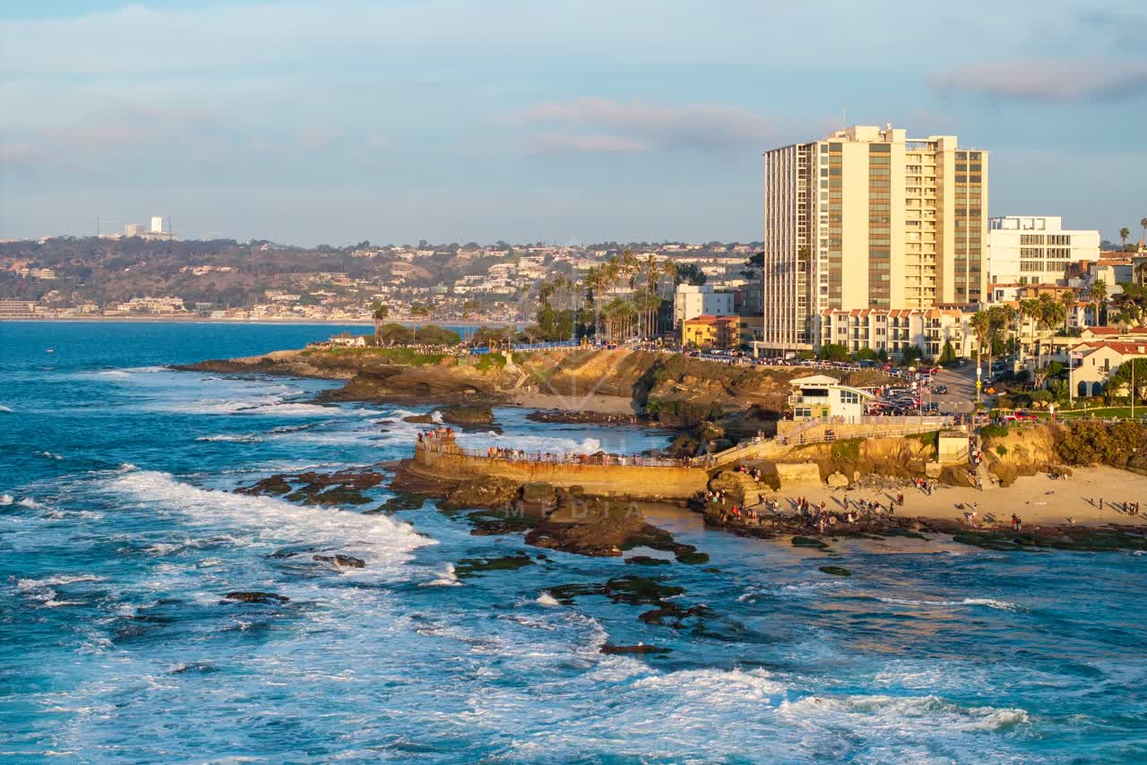 Low aerial view of the cliffs, the surf and palm trees at La Jolla Cove, with a crowd gathered.