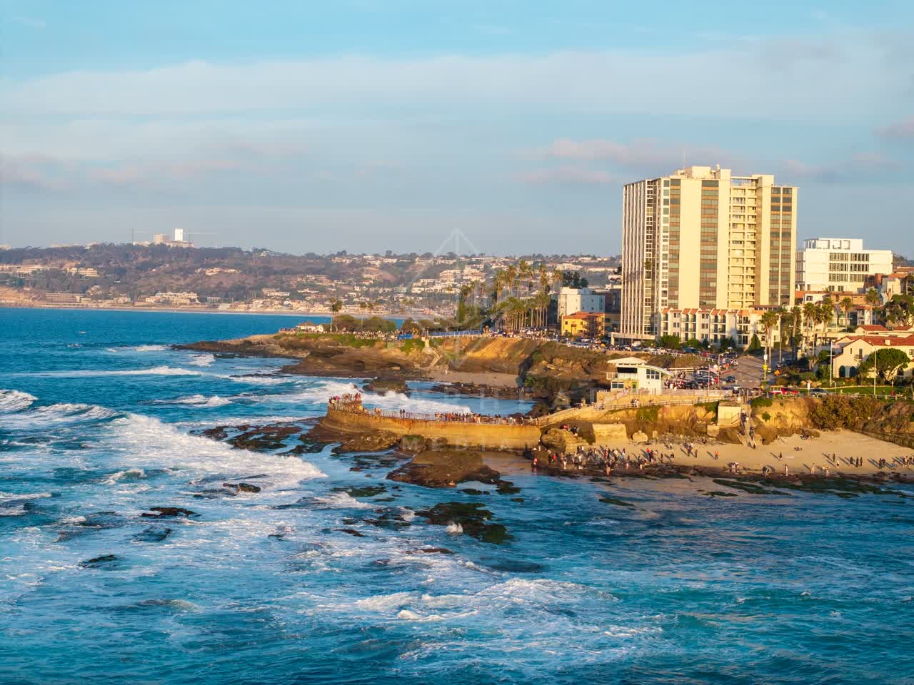 Aerial view of the cliffs, the surf and palm trees at La Jolla Cove, with a crowd gathered.