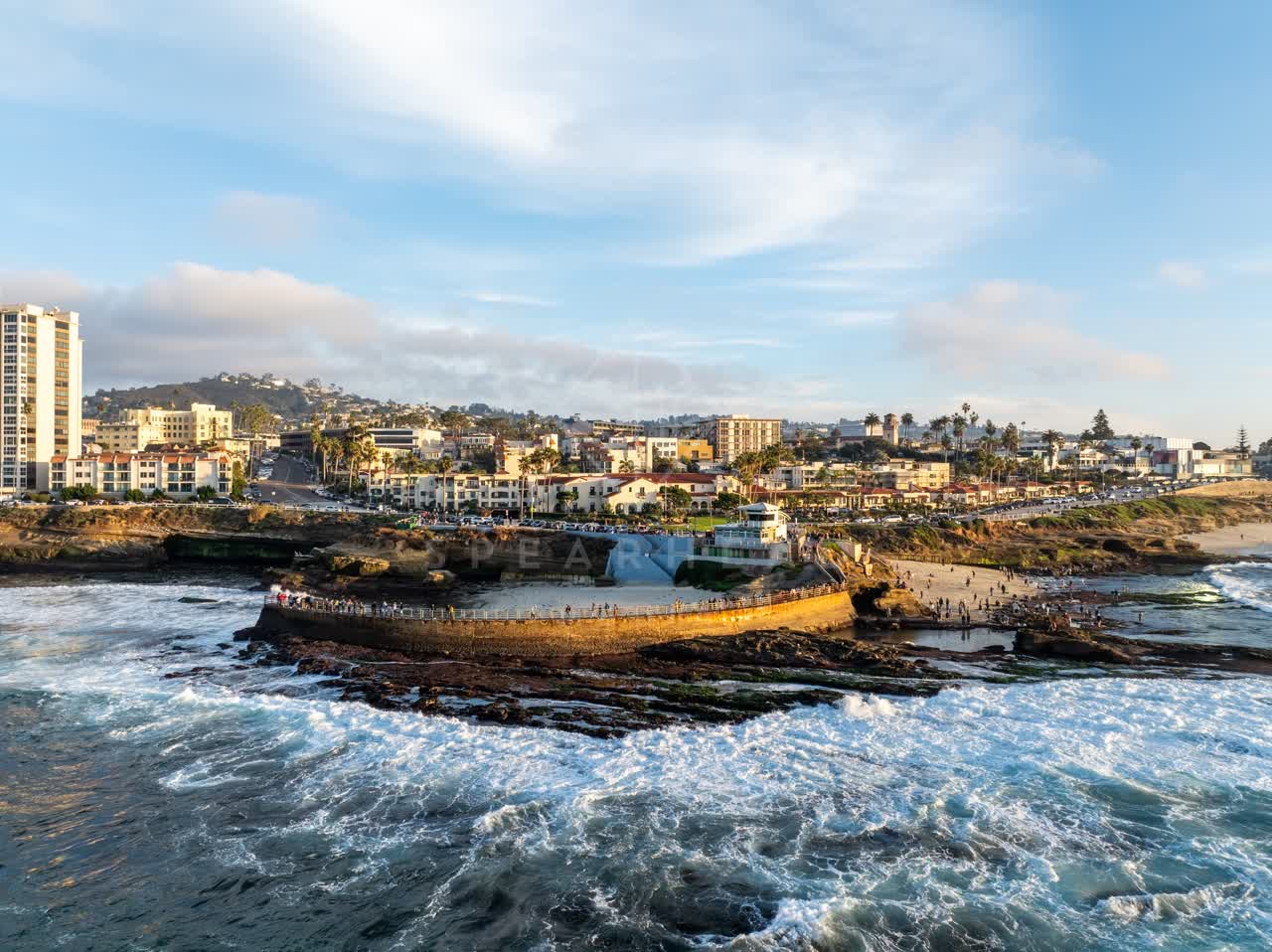 Aerial view of the cliffs, the surf and palm trees at La Jolla Cove, with a crowd gathered.
