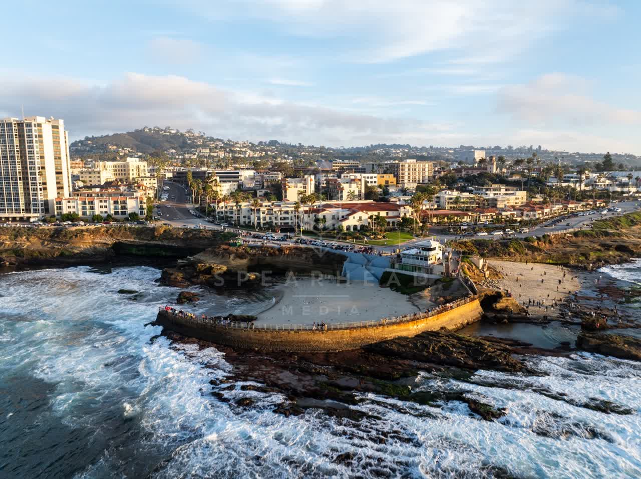 Aerial view of the cliffs, the surf and palm trees at La Jolla Cove, with a crowd gathered.