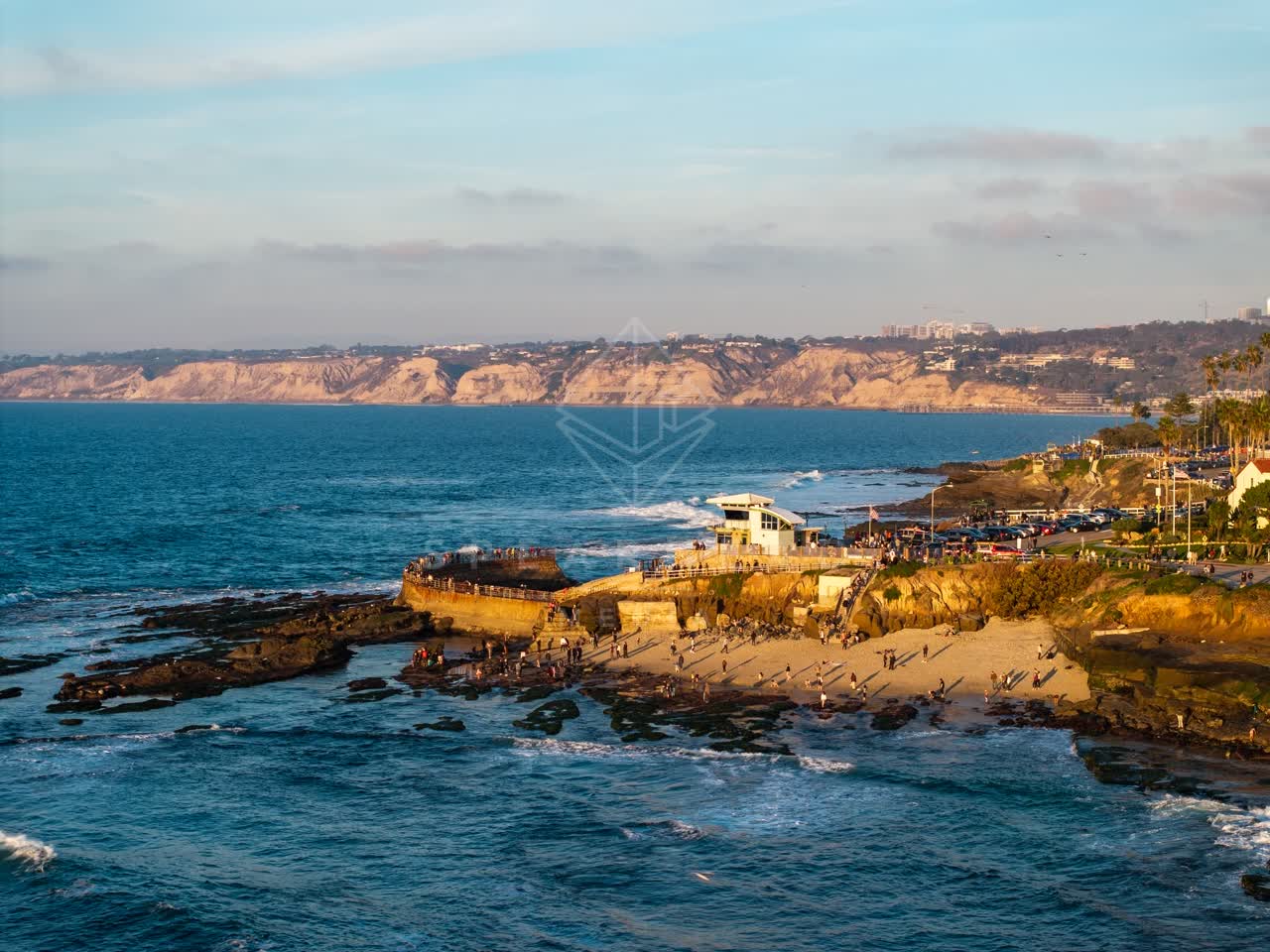 Aerial view of the cliffs, the surf and palm trees at La Jolla Cove, with a crowd gathered.