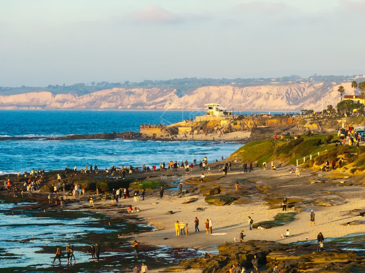 Low aerial view of the beach, the ocean and the cliffs at La Jolla Cove, with a crowd gathered.